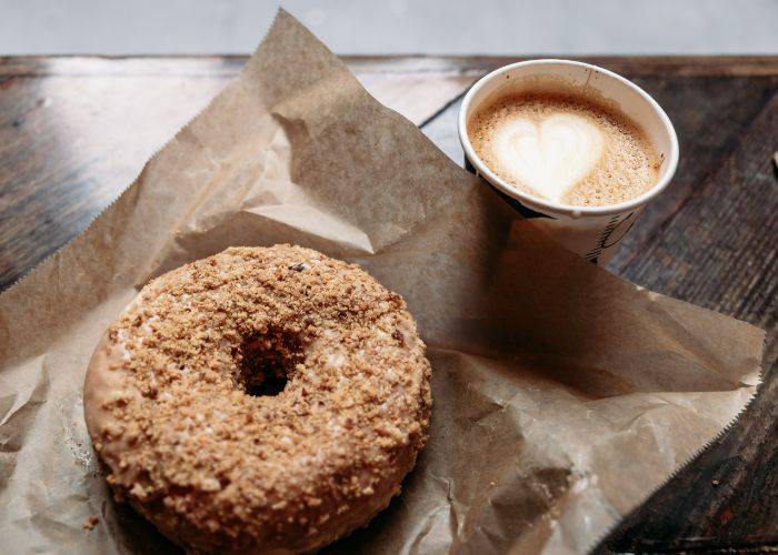 A donut with a biscuit crumb next to a coffee with heart in the foam.